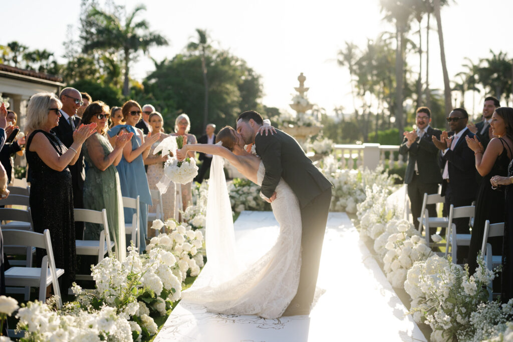 Bride and groom sharing a romantic kiss at the end of a flower-lined outdoor wedding aisle with guests applauding under palm trees at sunset