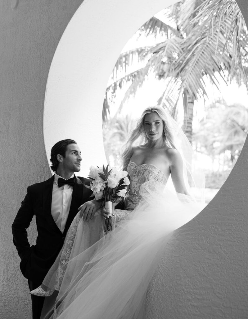 Black and white photo of a bride holding a bouquet, sitting gracefully near a circular architectural opening with the groom standing beside her in a tuxedo.