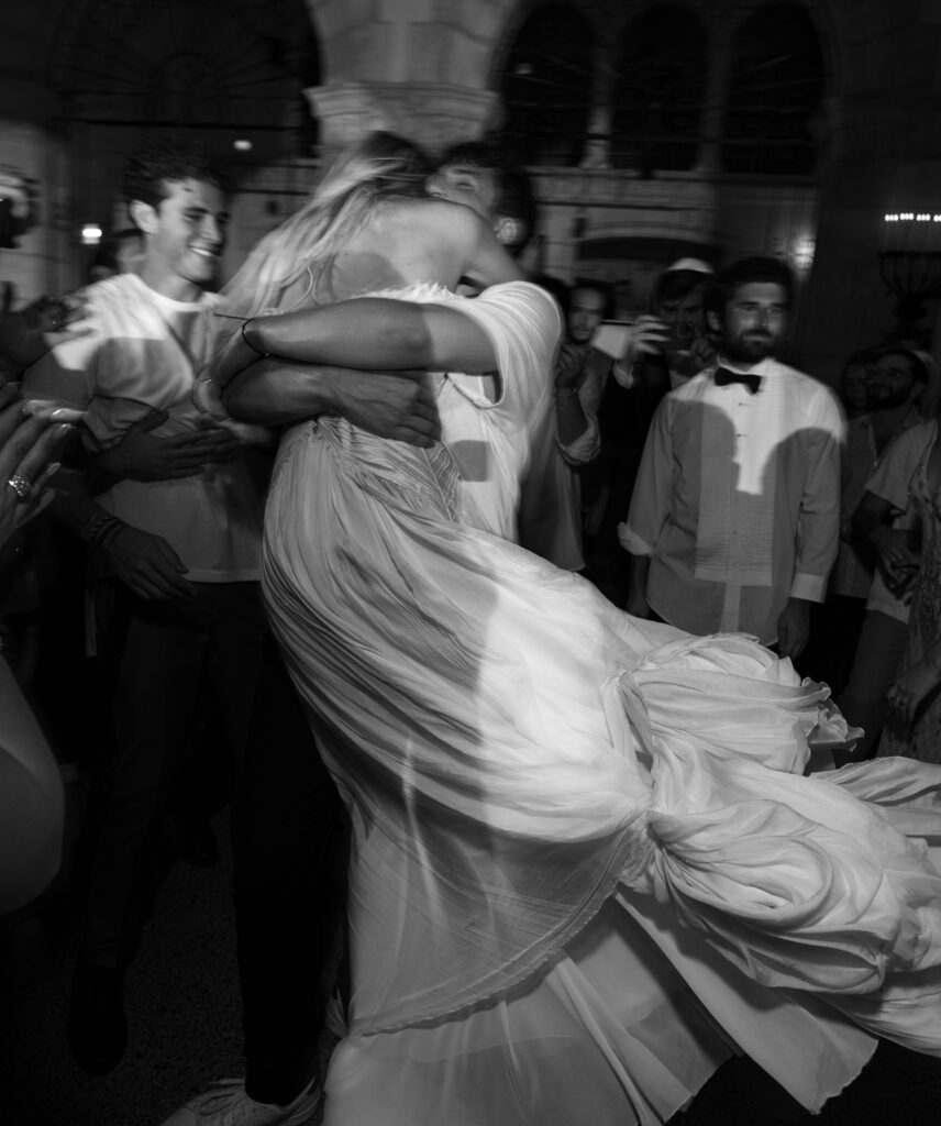 A romantic black-and-white photo of the groom lifting and embracing the bride on the dance floor, her dress swirling around them as guests cheer and smile in the background