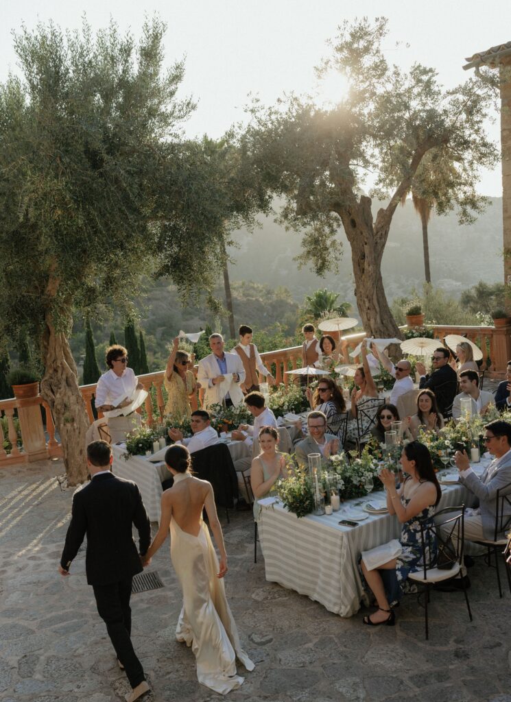 A couple, seen from behind, walks toward a stylish outdoor wedding reception with guests seated at tables decorated with flowers and greenery, surrounded by olive trees and warm sunlight.