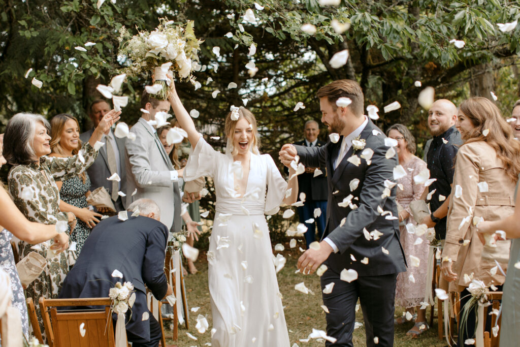 Bride and groom walking down the aisle as guests throw white flower petals at outdoor wedding ceremony.