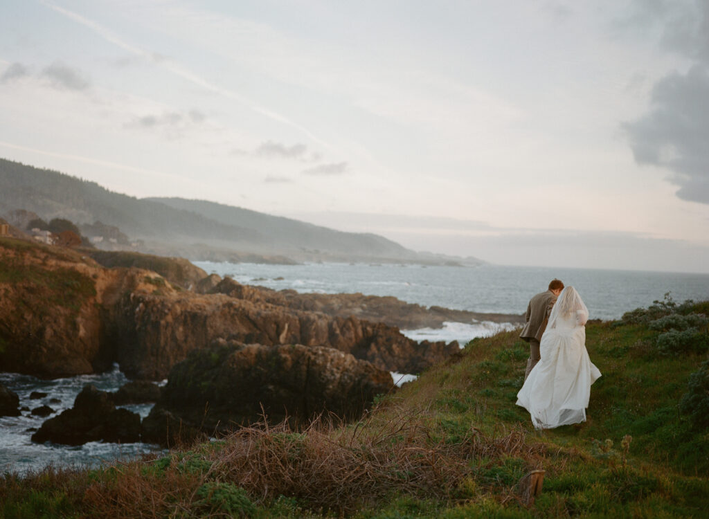  Bride and groom walking along a scenic coastal cliffside with ocean views on wedding day.